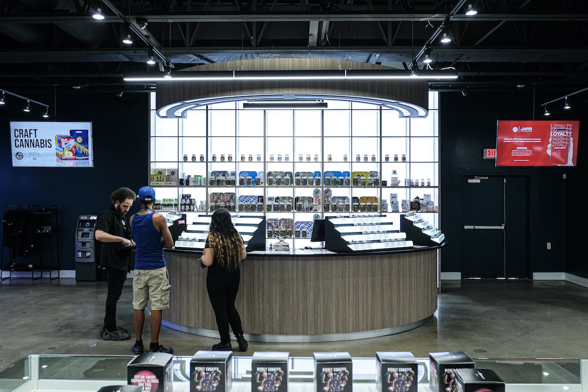 Two dispensary TV's on either side of the budwall. Shoppers sniff weed jars with promotions on the TVs