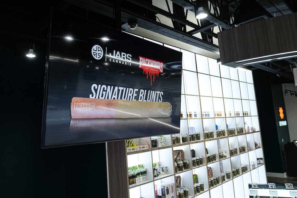 a TV hangs from the ceiling in JARS Cannabis (Michigan), displaying a promotional ad for Packwoods signature blunts